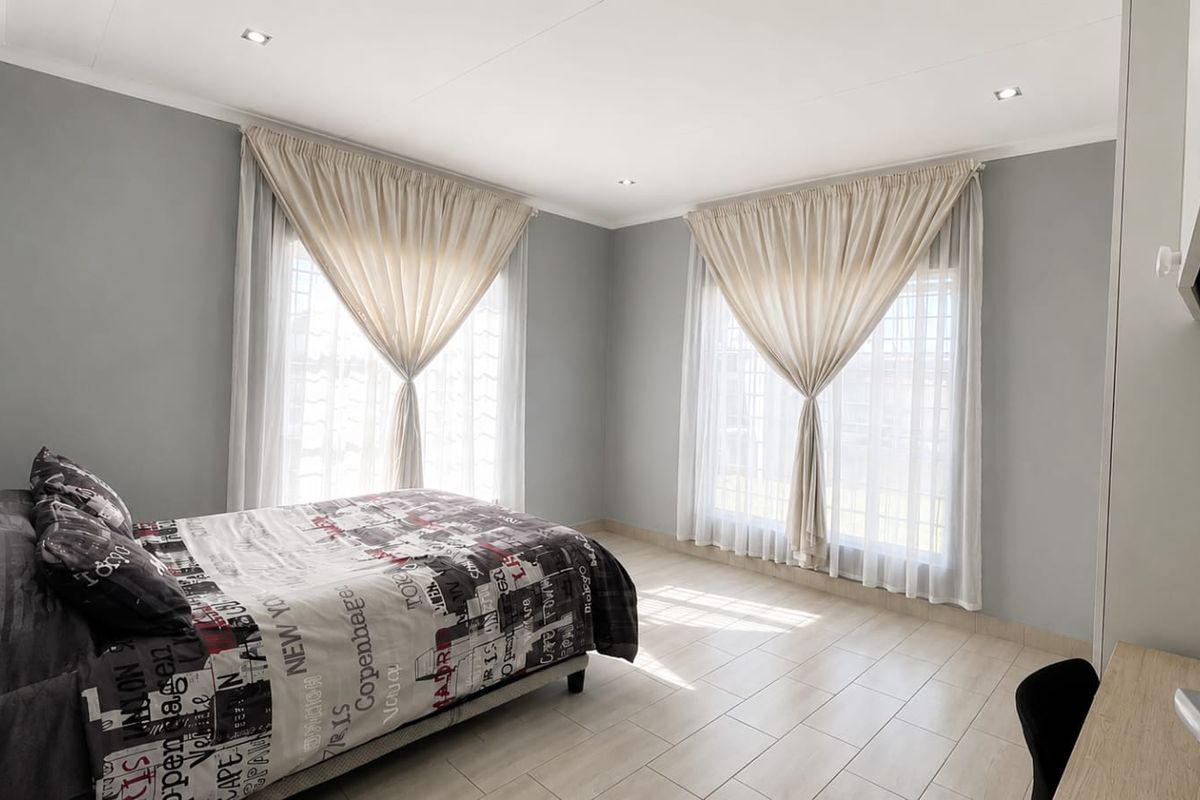 A bright and spacious bedroom featuring neutral grey walls, tiled flooring, and large windows with elegant drapery, creating a clean and comfortable living space.