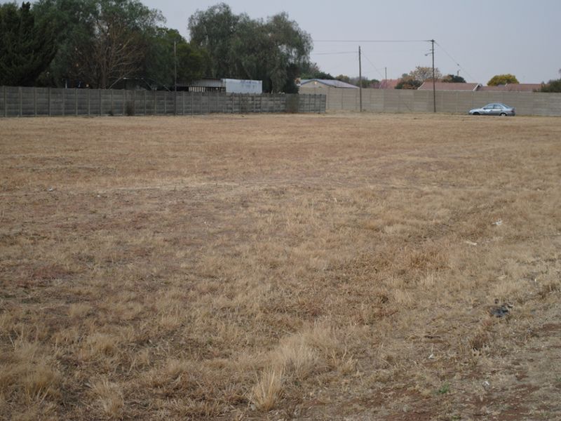 A vacant residential plot featuring dry grass, enclosed by a precast concrete boundary wall with overhead power lines visible in the background.