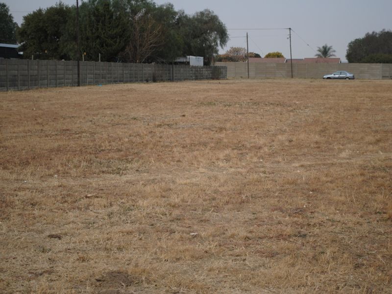 A vacant residential plot featuring dry grass, enclosed by a precast concrete boundary wall with visible utility infrastructure.