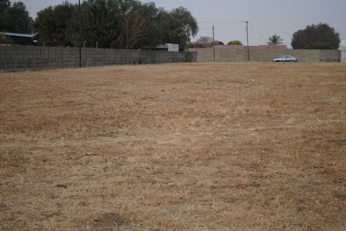 A vacant residential plot featuring dry grass, enclosed by a precast concrete boundary wall with visible utility infrastructure.