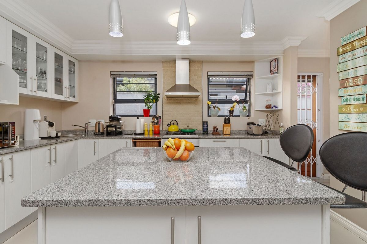A well-maintained kitchen featuring a large granite-topped island, ample white cabinetry, stainless steel extractor fan, and modern pendant lighting.