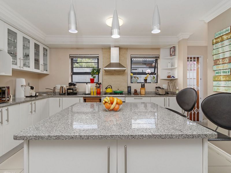 A well-maintained kitchen featuring a large granite-topped island, ample white cabinetry, stainless steel extractor fan, and modern pendant lighting.