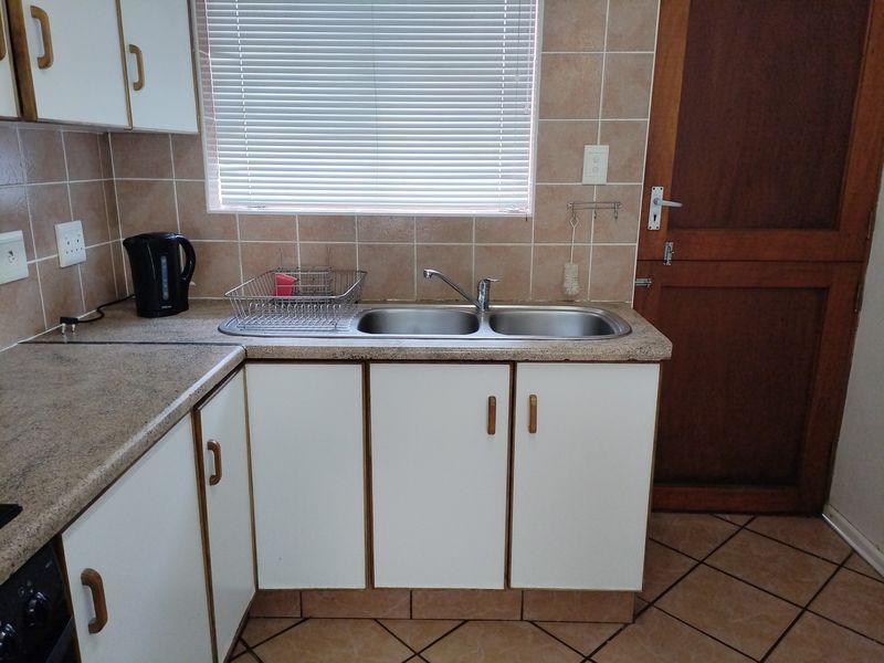A functional kitchen area featuring a double basin stainless steel sink, laminate countertops, tiled backsplash, and built-in cabinetry with tiled flooring.