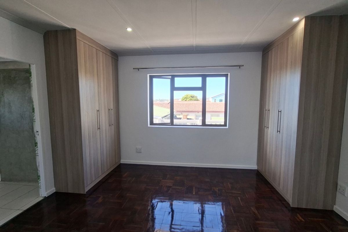 A bright, empty bedroom featuring polished parquet flooring, extensive built-in cupboards, and recessed lighting, with an adjoining bathroom visible.