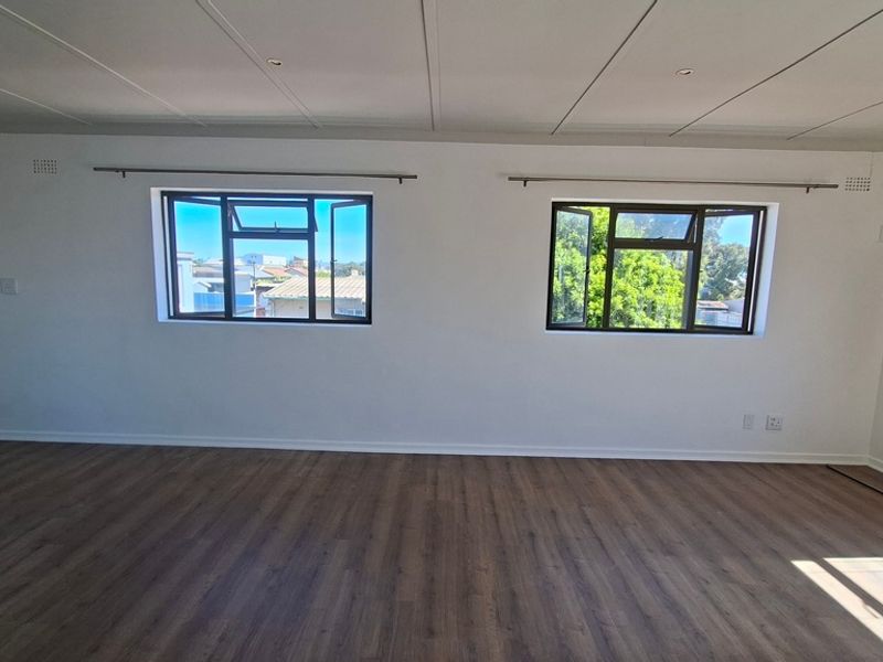 A bright, empty living room featuring modern laminate flooring, recessed ceiling lighting, and dual casement windows providing natural light.