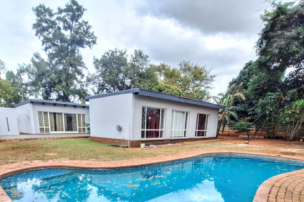 Rear view of the property featuring a swimming pool with brick coping, set against a garden with mature trees and a paved patio area.