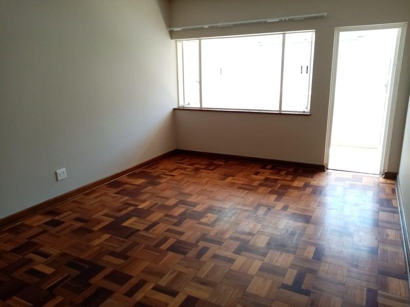 An empty bedroom featuring classic parquet flooring, neutral walls, and natural light from a large window and adjacent door.