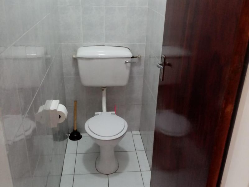 A compact, functional bathroom featuring white ceramic wall and floor tiles, a standard toilet, and a small window for ventilation.