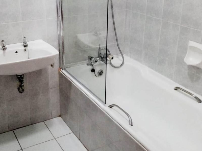 A functional bathroom featuring a wall-mounted basin, a bathtub with a glass shower screen, and neutral ceramic tiling throughout.