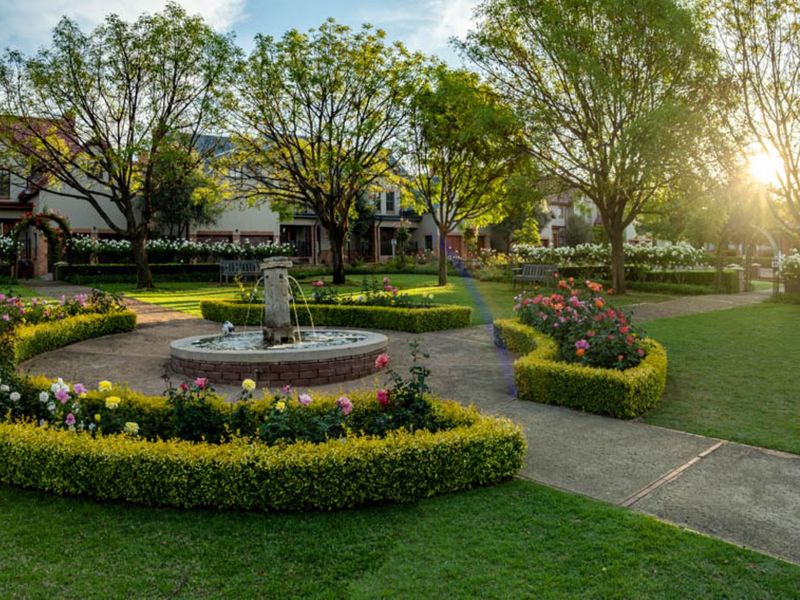 A beautifully landscaped communal garden featuring a central stone fountain, manicured hedges, mature trees, and paved walkways.