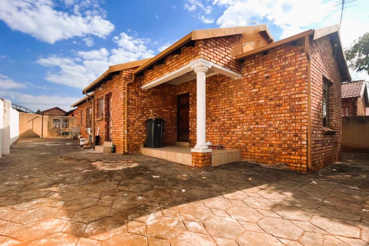 A side view of a face-brick house featuring a covered entrance porch with a decorative column and extensive paved outdoor area.