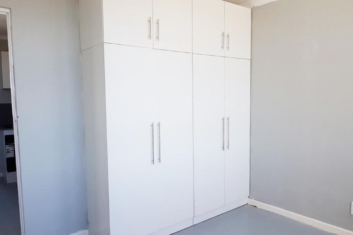 A clean, empty bedroom featuring floor-to-ceiling white built-in cupboards with modern handles and neutral grey wall finishes.