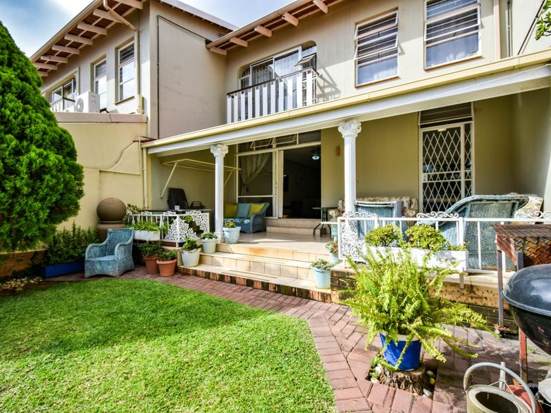 Rear view of the townhouse featuring a covered patio with decorative columns, tiled flooring, and a small balcony above, overlooking a neat garden.