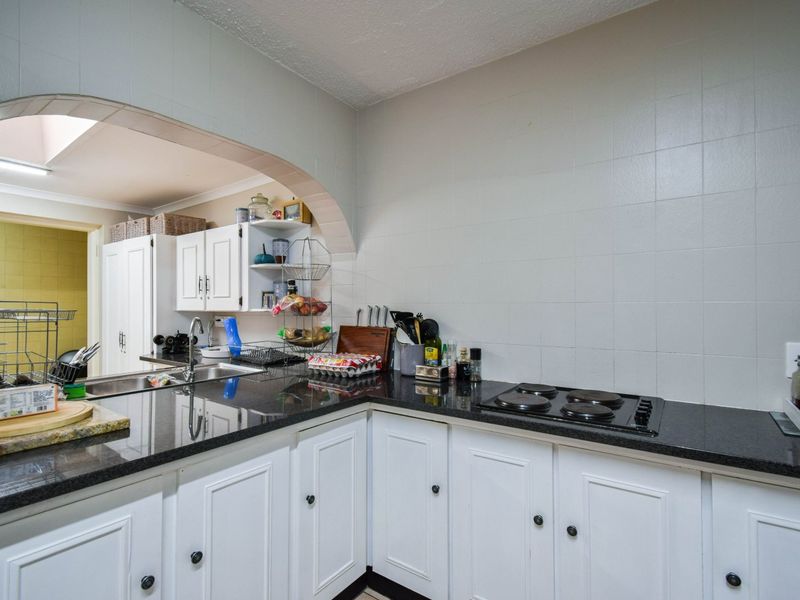 A functional kitchen featuring white cabinetry, dark granite countertops, an electric hob, and a tiled splashback, connected to an adjacent space via an arched opening.