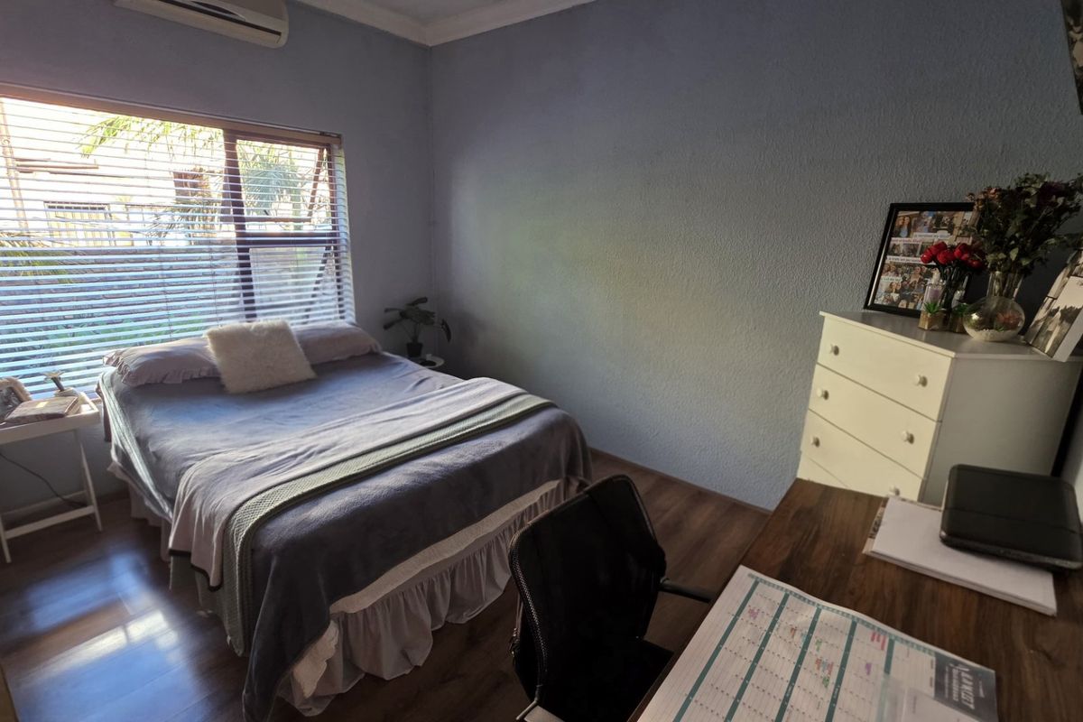 A functional bedroom featuring laminate flooring, a wall-mounted air conditioning unit, and natural light through venetian blinds.