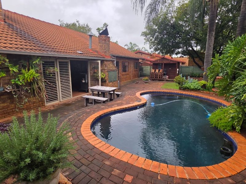 A rear garden area featuring a kidney-shaped swimming pool with brick coping, surrounded by paved patio space and established landscaping.