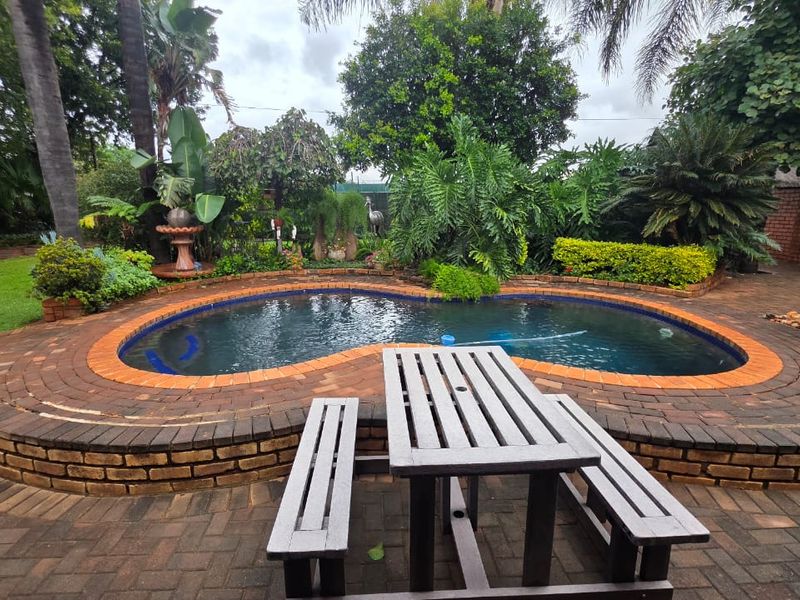 A well-maintained outdoor pool area featuring a kidney-shaped swimming pool with brick paving and established garden landscaping.