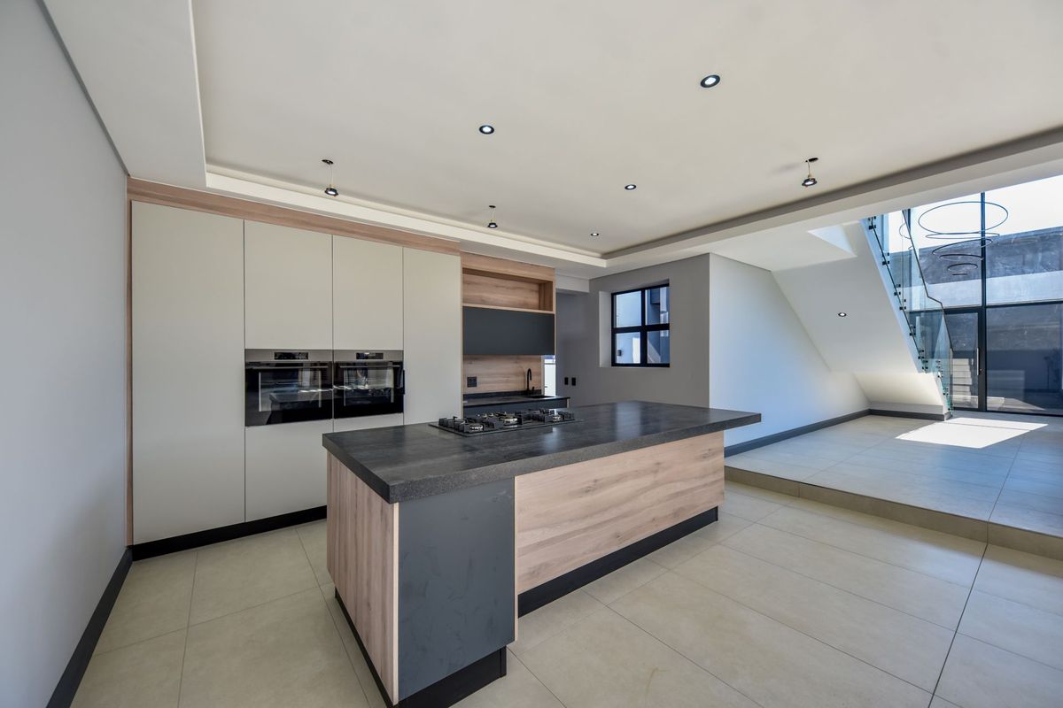 Modern kitchen featuring a central island with gas cooktop, built-in double ovens, sleek cabinetry, and tiled flooring, adjacent to a staircase with glass balustrade.