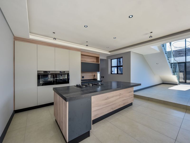 Modern kitchen featuring a central island with gas cooktop, built-in double ovens, sleek cabinetry, and tiled flooring, adjacent to a staircase with glass balustrade.