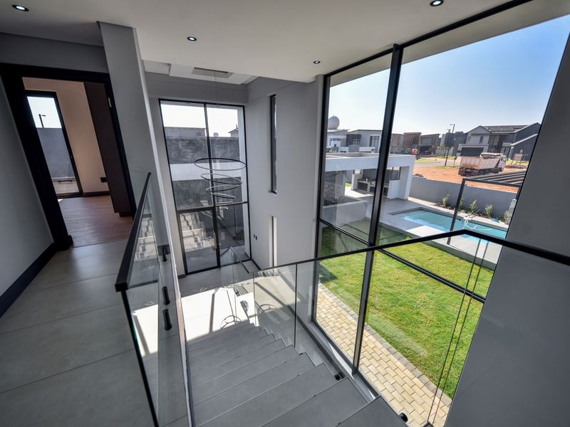 Modern double-volume hallway featuring a glass balustrade staircase, large format floor tiles, and expansive floor-to-ceiling windows overlooking the garden and pool area.