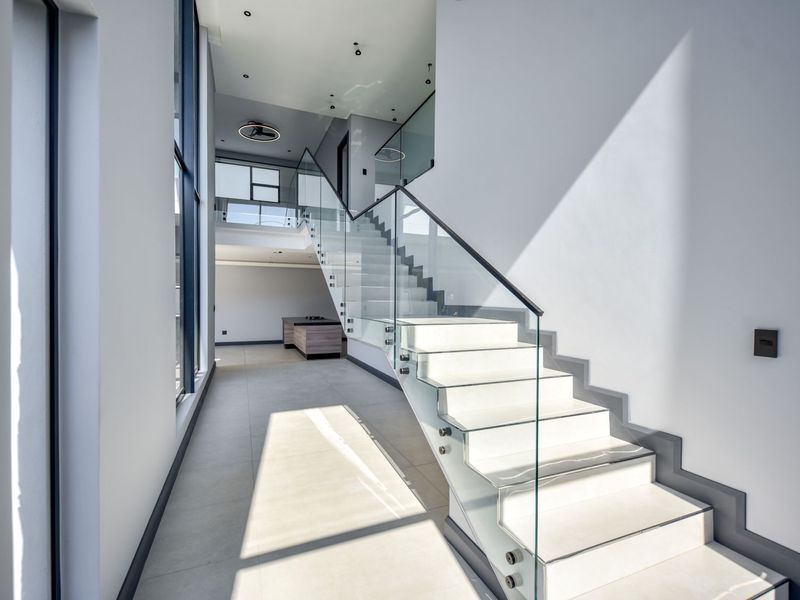 A modern, double-volume entrance hallway featuring a sleek glass-railed staircase and large-format floor tiles, creating a bright and spacious architectural aesthetic.