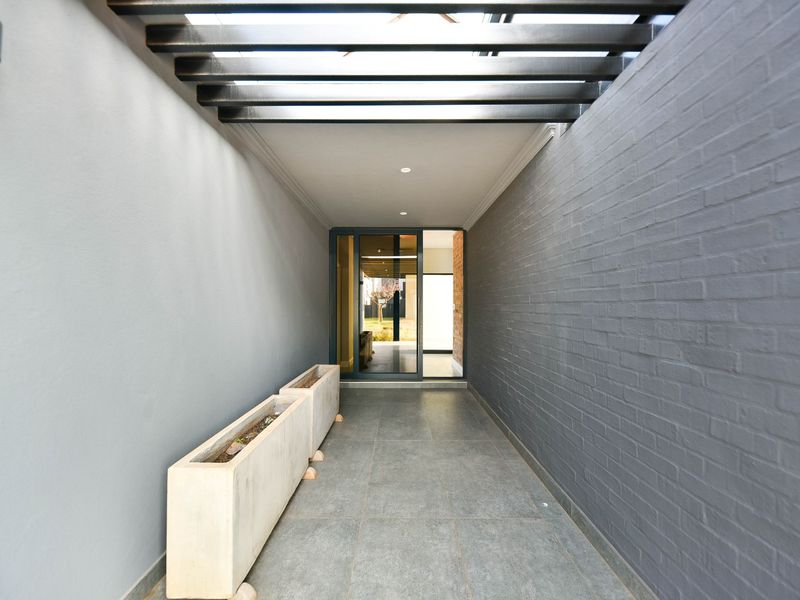 A modern, covered outdoor walkway or patio featuring a sleek pergola, grey tiled flooring, and a contemporary brick feature wall leading to a glass entrance.