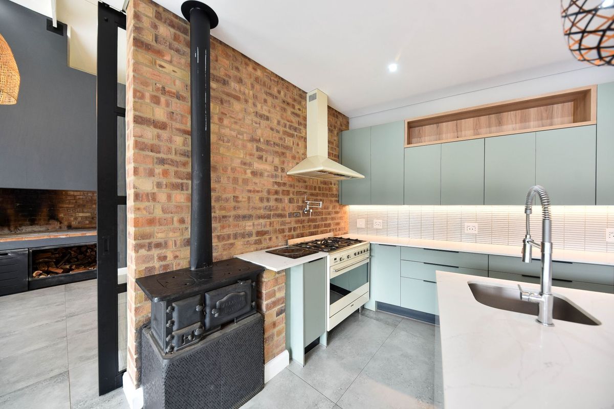 Modern kitchen featuring sage green cabinetry, stone countertops, a gas stove, and a striking exposed brick wall with a built-in wood-burning stove.