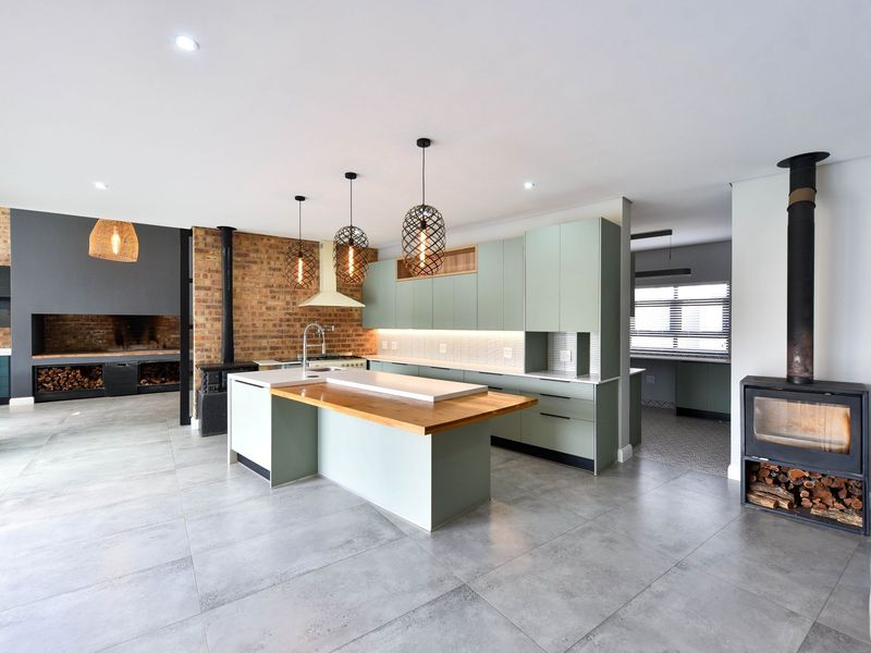 Modern open-plan kitchen featuring a central island, sage green cabinetry, exposed brick accents, and a cozy wood-burning fireplace in a spacious living area.