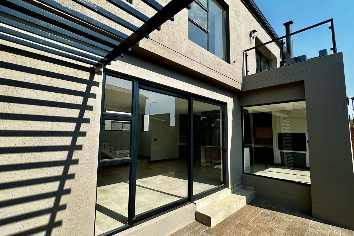 Rear view of a modern home featuring a paved patio, metal pergola, large sliding glass doors, and an upper-level balcony with a glass balustrade.