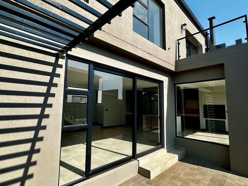 Rear view of a modern home featuring a paved patio, metal pergola, large sliding glass doors, and an upper-level balcony with a glass balustrade.