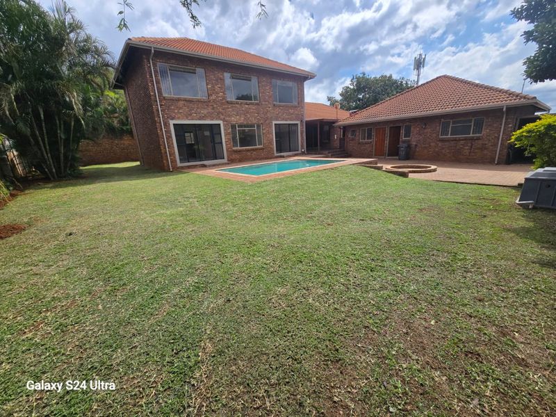 Rear view of a double-story brick house featuring a swimming pool, paved patio area, and a built-in fire pit set within a spacious garden.