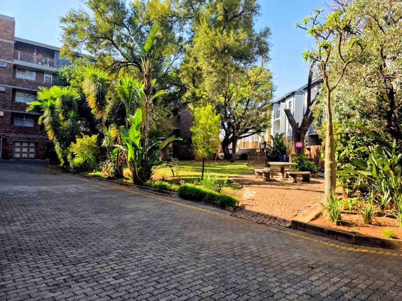 A well-maintained communal garden area featuring mature trees, landscaped greenery, and a paved driveway within an apartment complex setting.