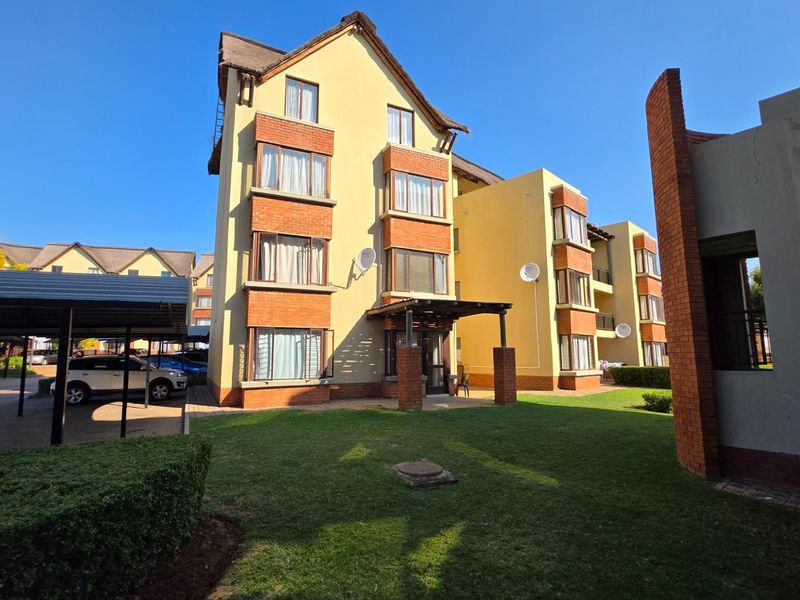 Front view of a multi-story apartment complex featuring brick and plaster facade, covered parking bays, and landscaped lawn area.