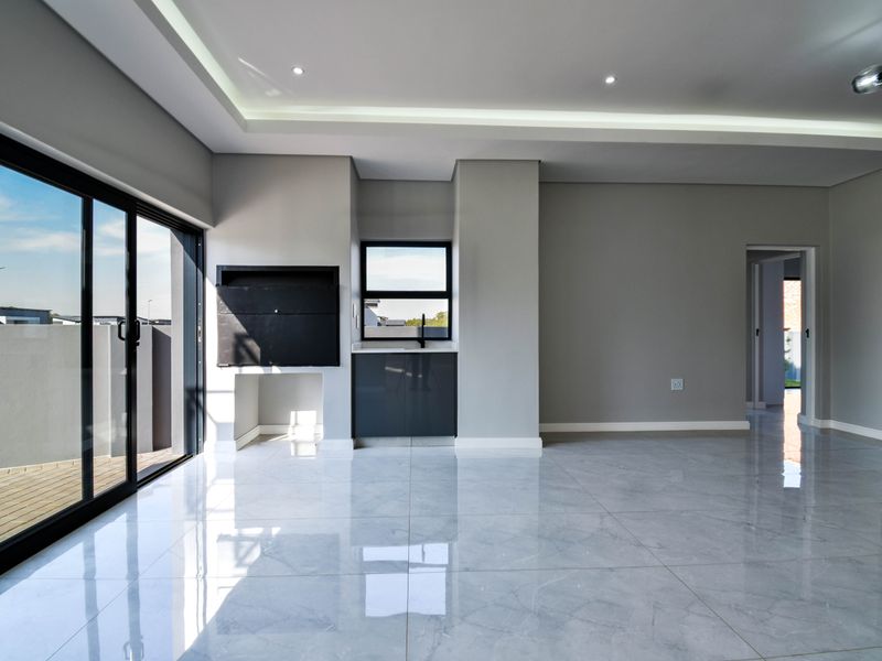 Modern open-plan living area featuring large sliding doors, polished tiled floors, recessed ceiling lighting, and a built-in braai with a small kitchenette area.