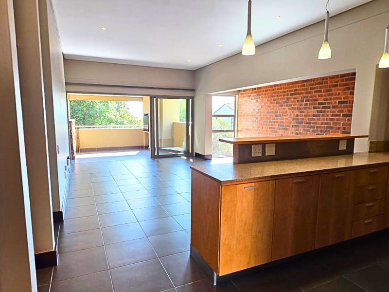 Spacious open-plan living area featuring a modern kitchen with stone countertops, breakfast bar, exposed brick wall, and seamless flow to a covered balcony.