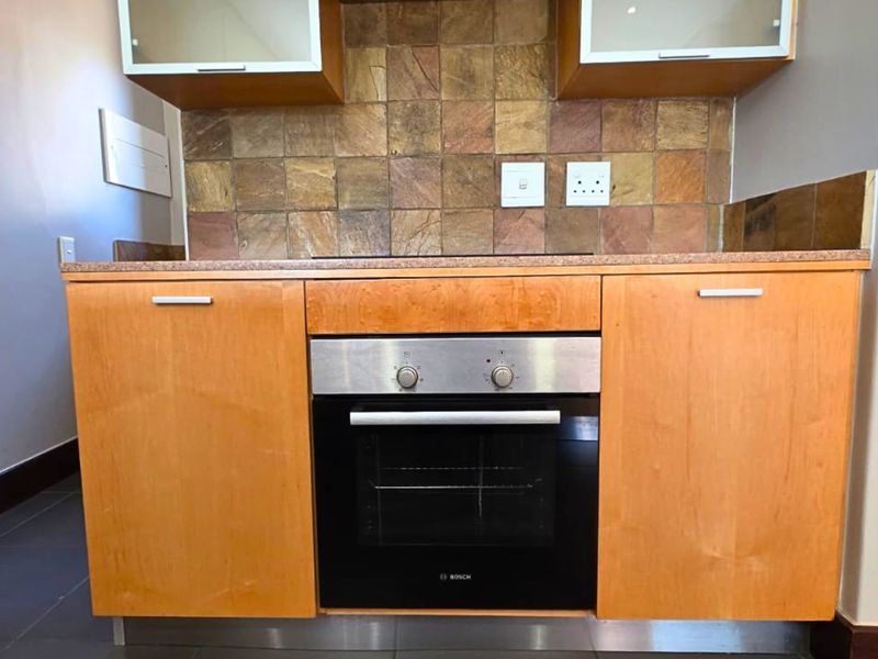 A compact kitchen area featuring wooden cabinetry, a built-in oven, and a textured stone tile backsplash.