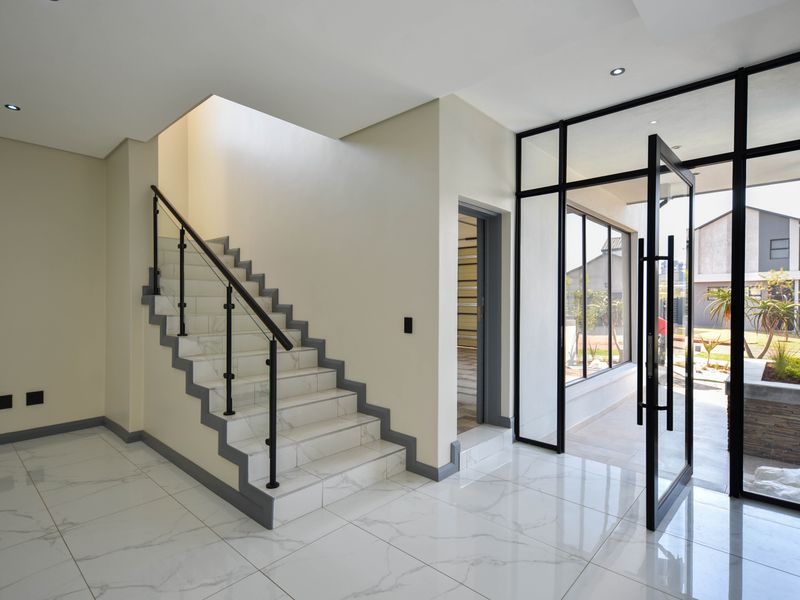 Modern entrance hall featuring large format marble-look floor tiles, a sleek staircase with metal balustrade, and a striking black-framed glass pivot door.