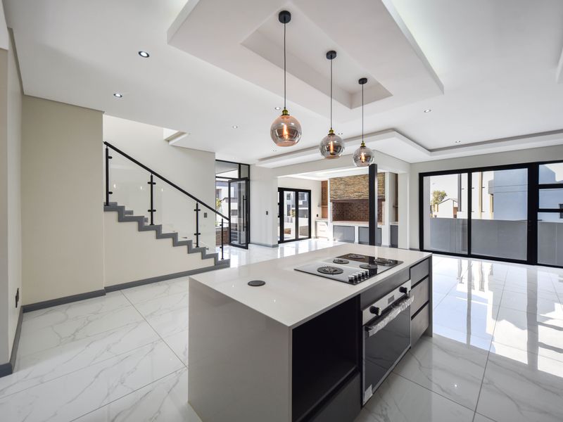 Modern open-plan living area featuring a sleek kitchen island with built-in appliances, marble-look flooring, recessed ceiling details, and a glass-balustrade staircase.