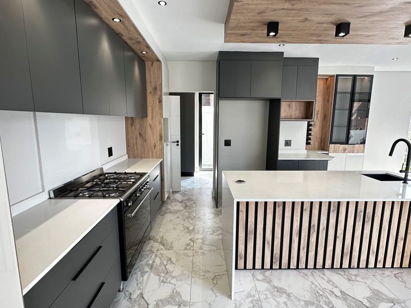 Modern kitchen featuring dark cabinetry, wood-look accents, stone countertops, a central island with a sink, and high-end built-in appliances.