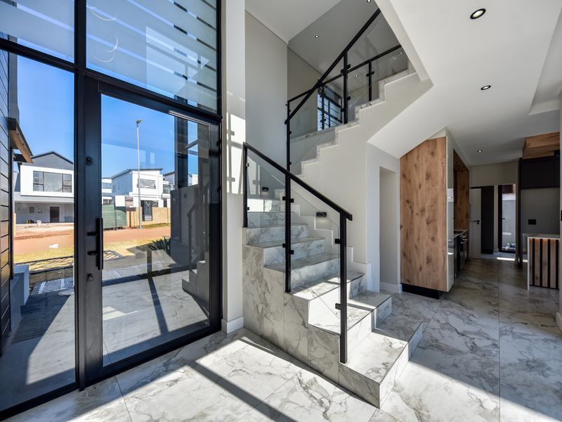 Modern double-volume entrance hall featuring marble-look tiled flooring, a sleek glass staircase balustrade, and expansive glass doors providing abundant natural light.