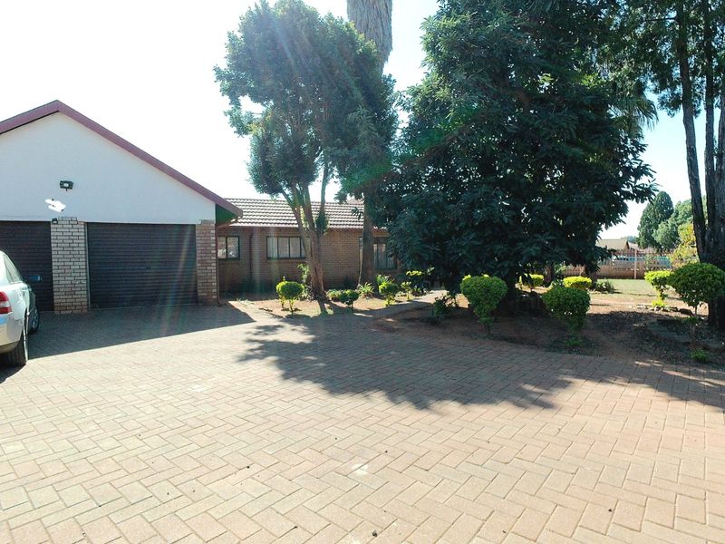 Front view of a single-story facebrick house featuring a paved driveway, double garage with roller doors, and established garden trees.