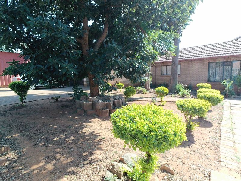 A front garden area featuring established trees, manicured shrubs, and a brick-faced house with a tiled roof and security bars on the windows.
