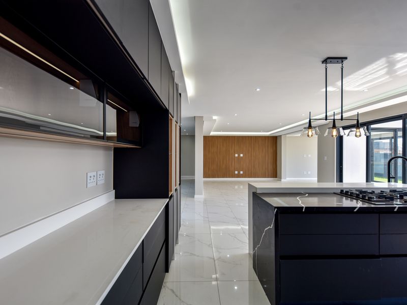 Modern open-plan kitchen and living area featuring sleek dark cabinetry, stone countertops, a central island with gas hob, and polished porcelain tile flooring.