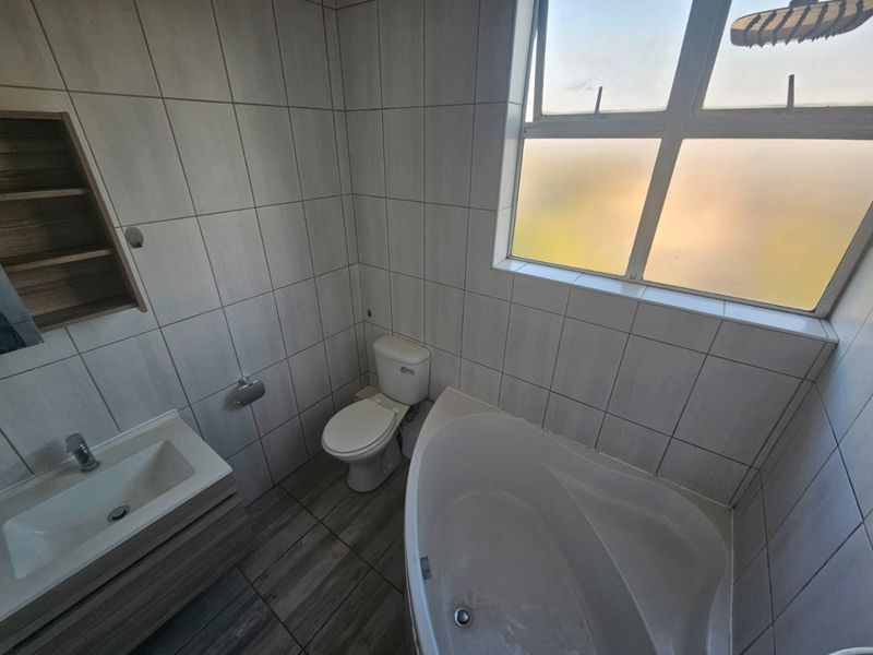 A clean, functional bathroom featuring a corner bathtub, vanity with basin, toilet, and neutral wall tiling with a frosted window for natural light.
