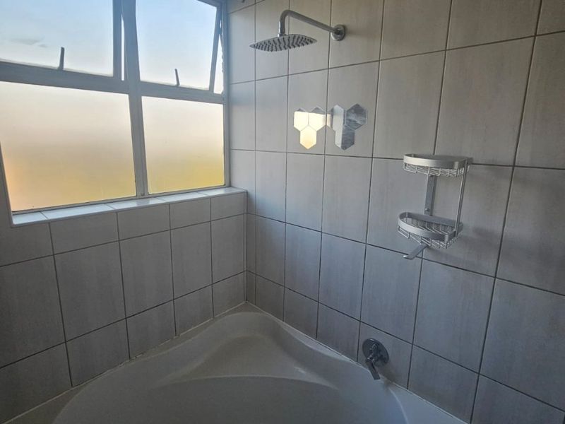 A bathroom featuring a corner bathtub with a wall-mounted shower head and tiled wall finishes, illuminated by a large window.