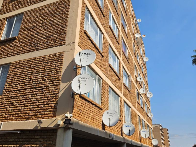 Exterior view of a multi-story brick apartment building featuring numerous satellite dishes mounted on the facade and a covered ground-level parking area.