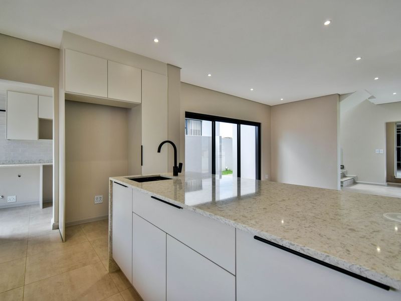 Modern kitchen featuring a central island with stone countertops, black fixtures, and sleek built-in cabinetry, leading to an adjacent scullery and outdoor access.