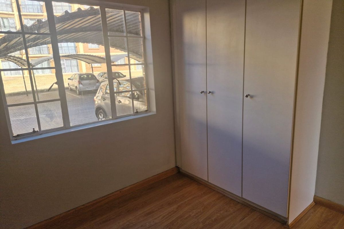 A bright, empty bedroom featuring laminate flooring and a built-in wardrobe with white doors, overlooking the apartment complex parking area.
