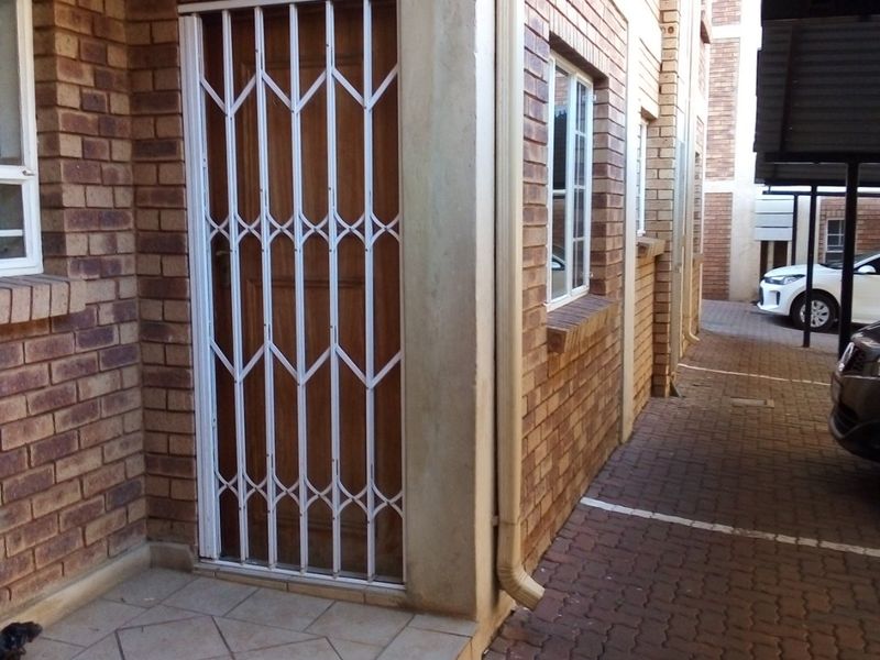 Exterior view of an apartment entrance featuring a face brick facade, security gate, tiled porch, and adjacent paved parking area with carport.