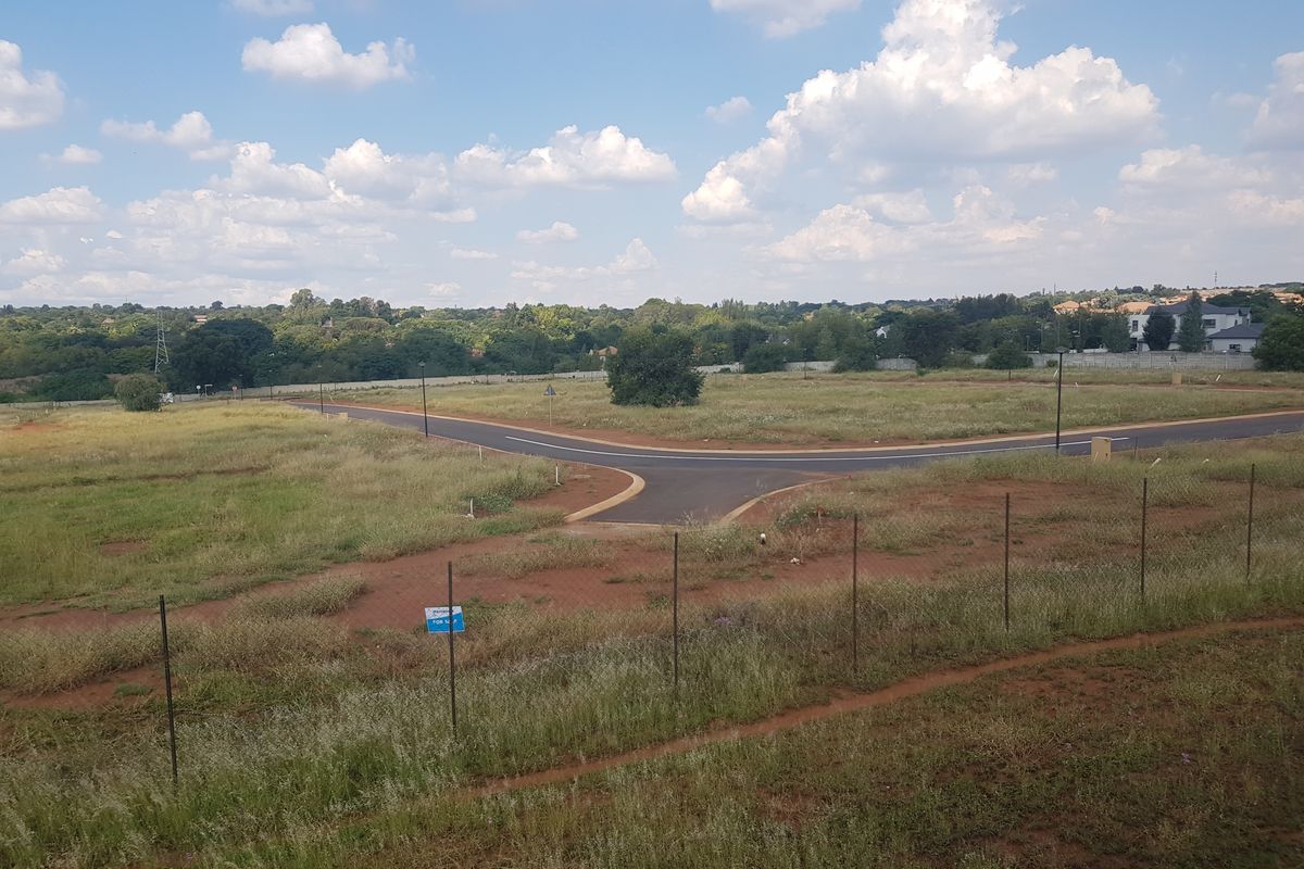 An elevated view of a vacant residential plot featuring established road infrastructure, perimeter fencing, and surrounding greenery in a developing estate.